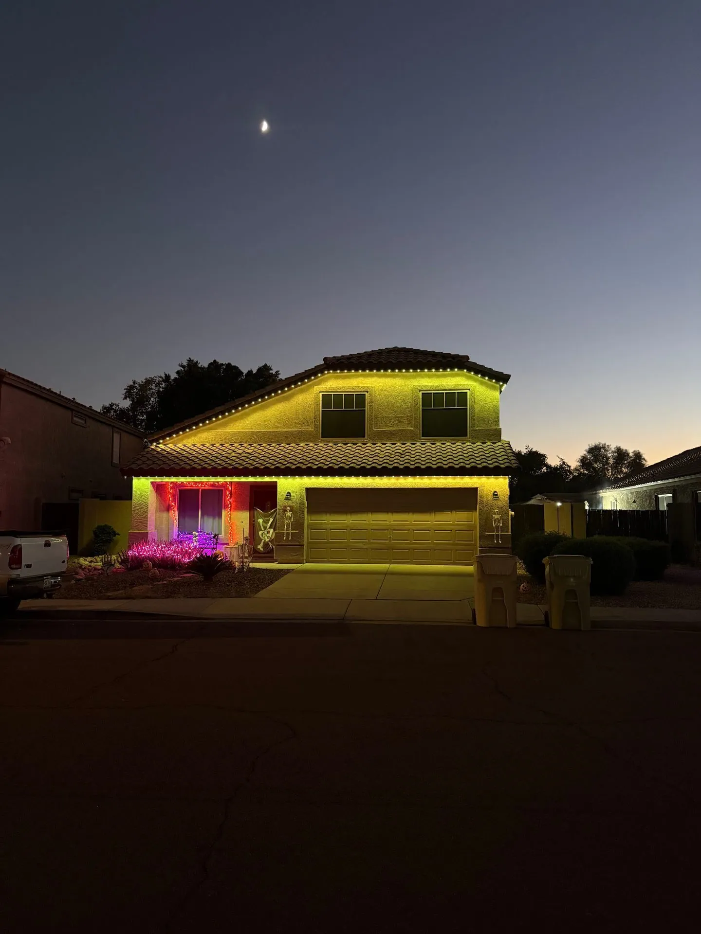 Multicolored roofline lights on a stucco home at dusk beneath a crescent moon